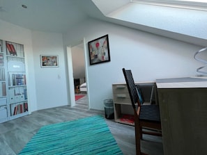 Ein Zimmer mit Oberlicht, einem Schreibtisch mit Stuhl, einem Bücherregal und einem gerahmten Bild an der Wand.