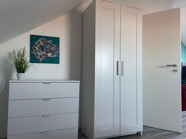 A room with a white wardrobe, a small white dresser, a potted plant, and a painting on the wall.