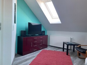 Ein Zimmer mit schrägen Wänden, einem Fernseher auf einem dunklen Schrank, einem weißen Heizkörper, einem kleinen Tisch mit Stuhl und einem roten Teppich.