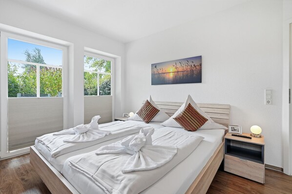 Une chambre d’hôtel avec un grand lit, deux fenêtres, un tableau, une table de chevet avec une lampe et une petite table de nuit.