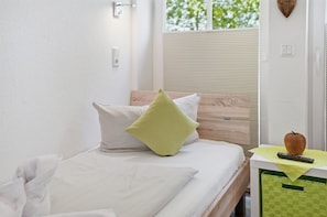 A bed with white bedding and two pillows, a wooden headboard, a remote control on a small table, and a green basket. - GrachtenEck - Bright vacation apartment in the canal district (Krummhörn)