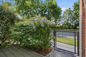 A gated entrance with a metal fence, a paved driveway, and a lush green hedge with purple flowers. - GrachtenEck - Bright vacation apartment in the canal district (Krummhörn)