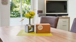 Une table en bois, sur laquelle se trouvent un vase de fleurs et une boîte en carton étiquetée « Pro-Uraub », le tout posé sur un set de table jaune.