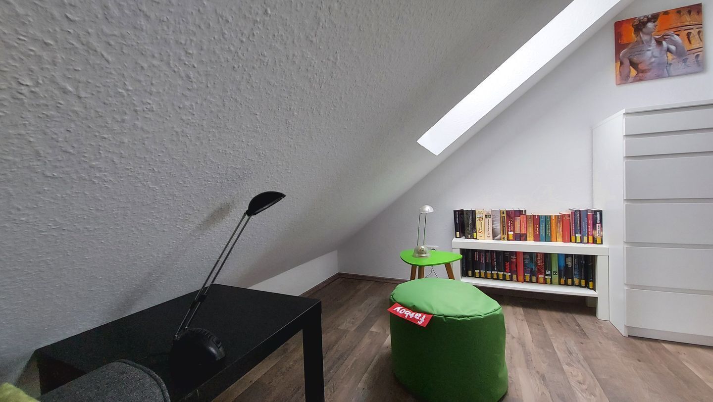Ein Zimmer mit schrägen Wänden, einem schwarzen Tisch mit Lampe, einem grünen Pouf, einem kleinen grünen Tisch und einem Bücherregal mit Büchern.