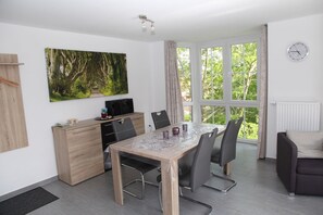 A modern dining area with a wooden table, black chairs, a black cabinet, and a large window with a view of greenery. - Kyana - Schöne Ferienwohnung in Ruhiger Lage von Norden (Norden)