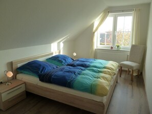 A bedroom with a wooden bed, a bedside table with a lamp, a chair, and a window with curtains. - Kyana - Schöne Ferienwohnung in Ruhiger Lage von Norden (Norden)