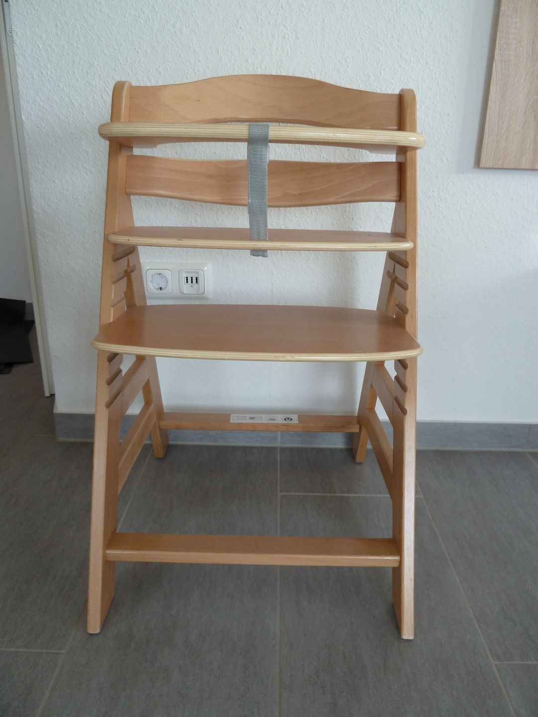 A wooden desk with three shelves, a grey strap, and a power outlet on the wall behind it.