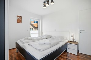 Ein ordentlich bezogenes Bett mit weißen Leinen, ein Nachttisch mit Lampe, eine kleine Kommode und ein Fenster mit Blick auf Gebäude.
