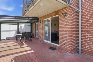 Une terrasse dotée d’une véranda en verre, de murs en briques et d’une porte-fenêtre coulissante donnant sur un espace intérieur.