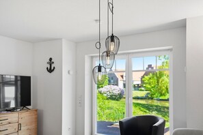 Un moderno salón con televisor, mueble de madera y vistas a un jardín a través de amplios ventanales.