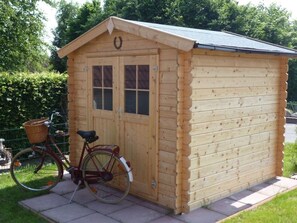 Un abri de jardin en bois, avec une porte et des fenêtres, un vélo garé devant et un panier sur le vélo.