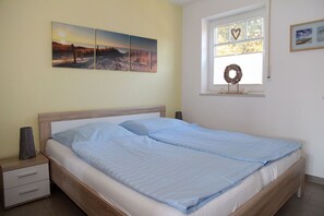 Un dormitorio con cama, mesita de noche, ventana adornada con un arreglo floral y pared con tres cuadros.