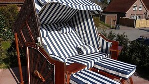 En randig utomhusfåtölj med ett solskydd, placerad på en stenlagd yta med ett hus och grönska i bakgrunden.