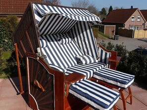 Una silla de exterior a rayas con toldo, ubicada en una zona pavimentada con una casa y vegetación al fondo.
