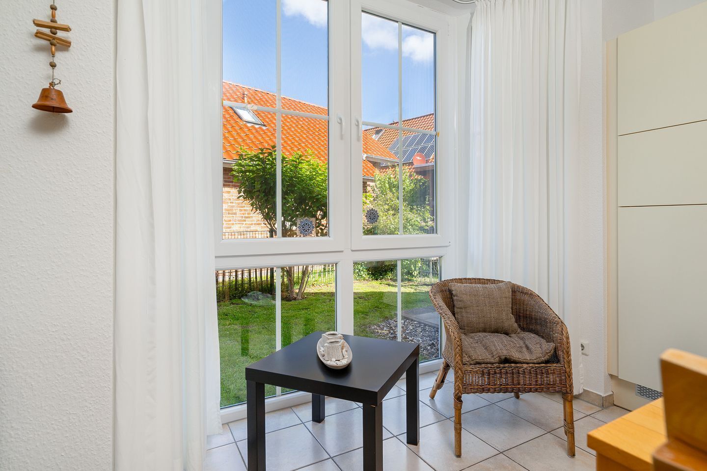A room with a large window, a wicker chair, a small table with a decorative item, and a white wardrobe.