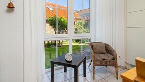 Una habitación con un ventanal grande, una silla de mimbre, una mesita con un objeto decorativo y un armario blanco.