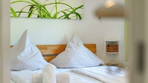 Una cama bien hecha con sábanas y almohadas blancas, un cabecero de madera y un pequeño estante con una cesta.