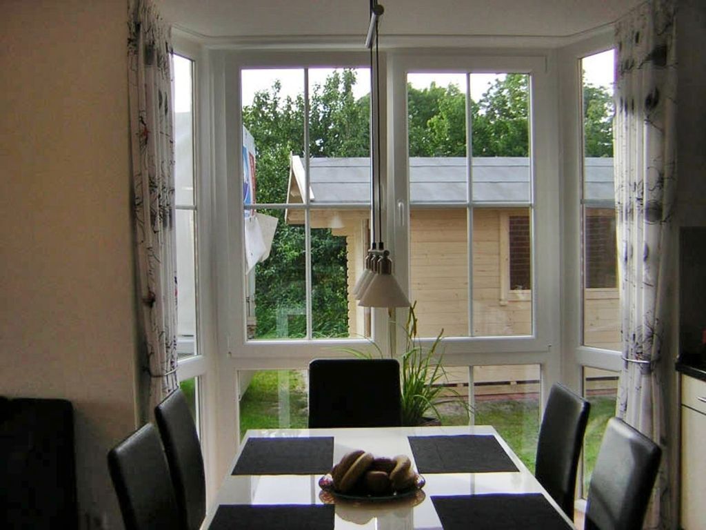 A dining area with a table and chairs, a window with a view of a house and greenery, and a hanging light fixture.