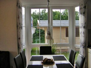 A dining area with a table and chairs, a window with a view of a house and greenery, and a hanging light fixture. - Gesa - Komfortable Ferienwohnung im Grachtenviertel (Krummhörn)