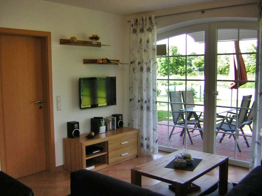 A living room with a flat-screen TV, wooden furniture, a coffee table, and a sliding glass door leading to an outdoor area with chairs.