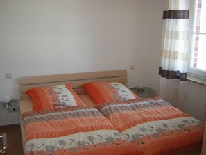 Un dormitorio con cabecera de madera, dos camas individuales con ropa de cama naranja y blanca, una mesita de noche con lámpara, y una ventana con cortinas.
