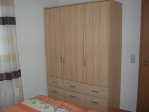 A bedroom with a wooden wardrobe, a bed with an orange and white bedspread, and a window with sheer curtains. - Gesa - Komfortable Ferienwohnung im Grachtenviertel (Krummhörn)