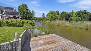 A wooden dock extending over a calm river, surrounded by lush greenery and a house with a sloped roof. - Friesenperle - Great vacation home (85sqm) for 4 people with fireplace directly on the (Krummhörn)