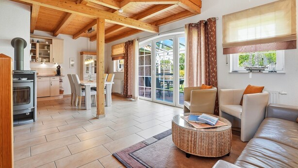 A spacious living area with a high ceiling, wooden beams, a dining area, and a view through large glass doors. - Friesenperle - Great vacation home (85sqm) for 4 people with fireplace directly on the (Krummhörn)