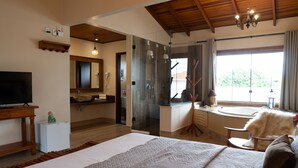A bedroom with a large bed, a television, a small refrigerator, a bathroom with a glass shower, and a view of the outdoors. - Pousada Lua Clara (Alto Paraíso de Goiás)