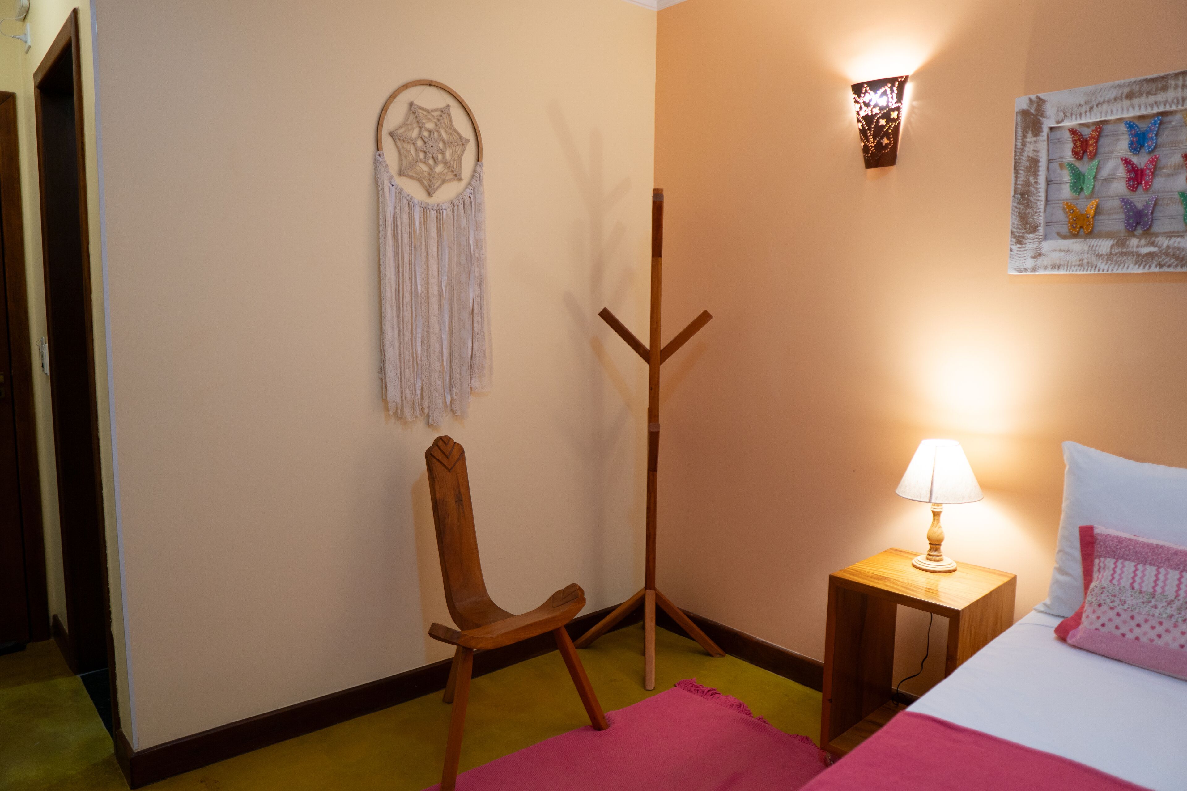 A bedroom with a bed, a nightstand with a lamp, a wooden chair, a dreamcatcher, and wall art.