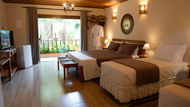 A hotel room with two beds, a TV, a desk, and a balcony with curtains. - Pousada Lua Clara (Alto Paraíso de Goiás)