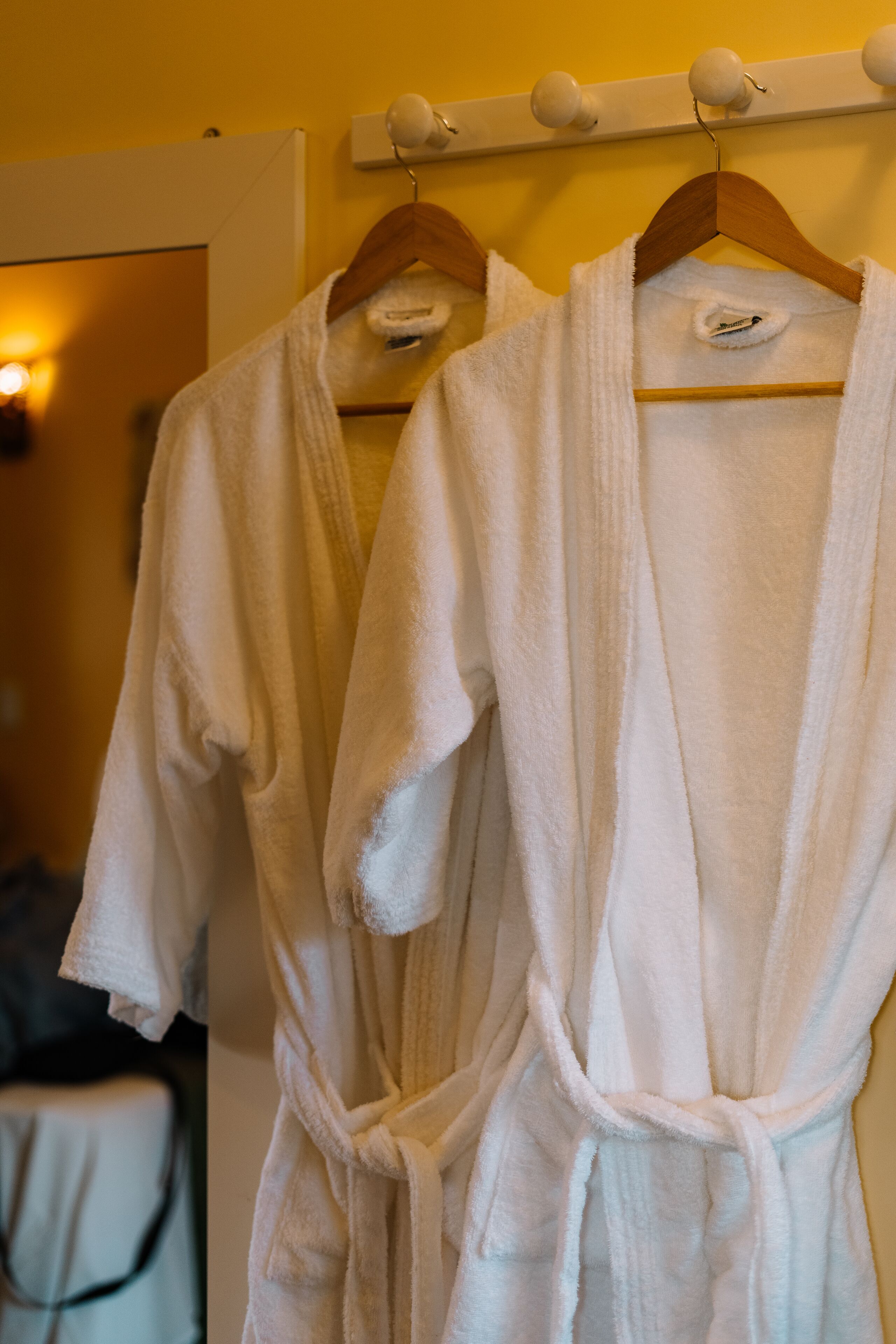 Two white bathrobes hanging on a wooden rack in a room with a yellow wall and a bed in the background.