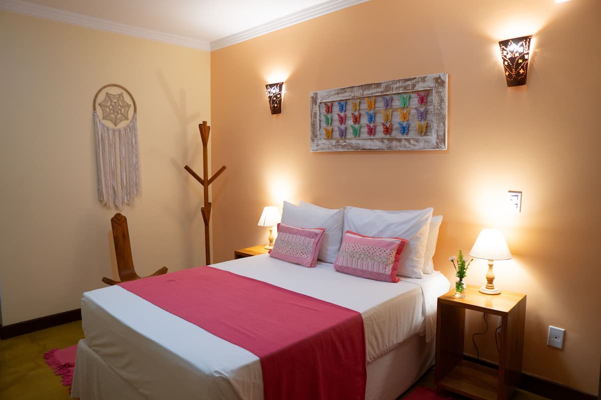 Cama bem arrumada com colcha rosa e almofadas brancas, um sonho capturado fixado na parede, arte de parede com borboleta colorida e duas luminárias de cabeceira.
