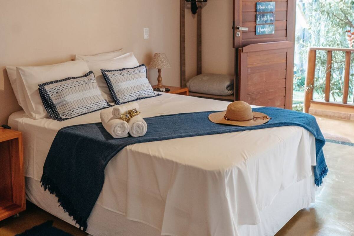 Quarto com cama, almofadas, cobertor, chapéu, toalha, mesa pequena, luminária, moldura de quadro, aparelho de ar condicionado, porta e vista para o verde lá fora.