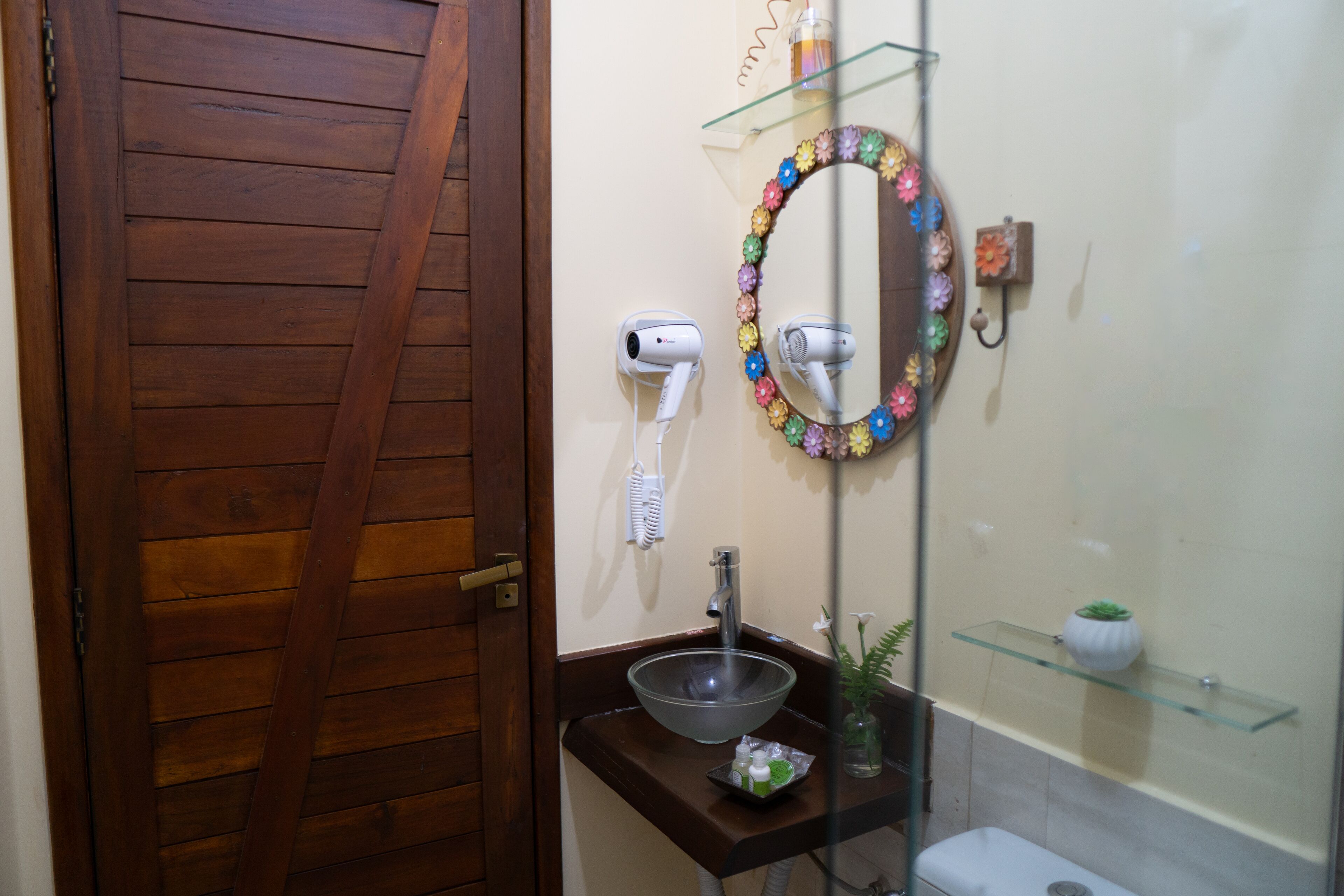 A bathroom with a wooden door, a glass sink, a mirror with a flower frame, and a hairdryer on the wall.