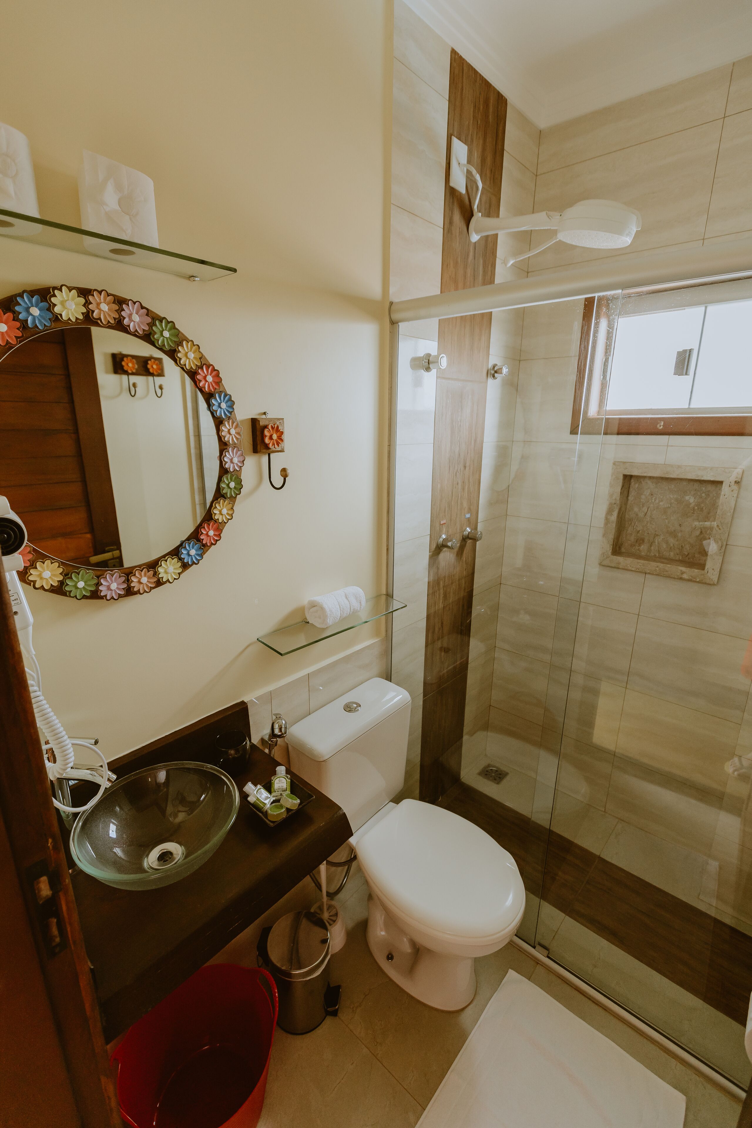 A bathroom with a glass shower enclosure, a toilet, a sink with a glass basin, and a mirror with a floral frame.