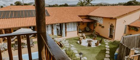A wooden balcony overlooking a courtyard with a swimming pool, stone pathway, and a fenced area with plants.