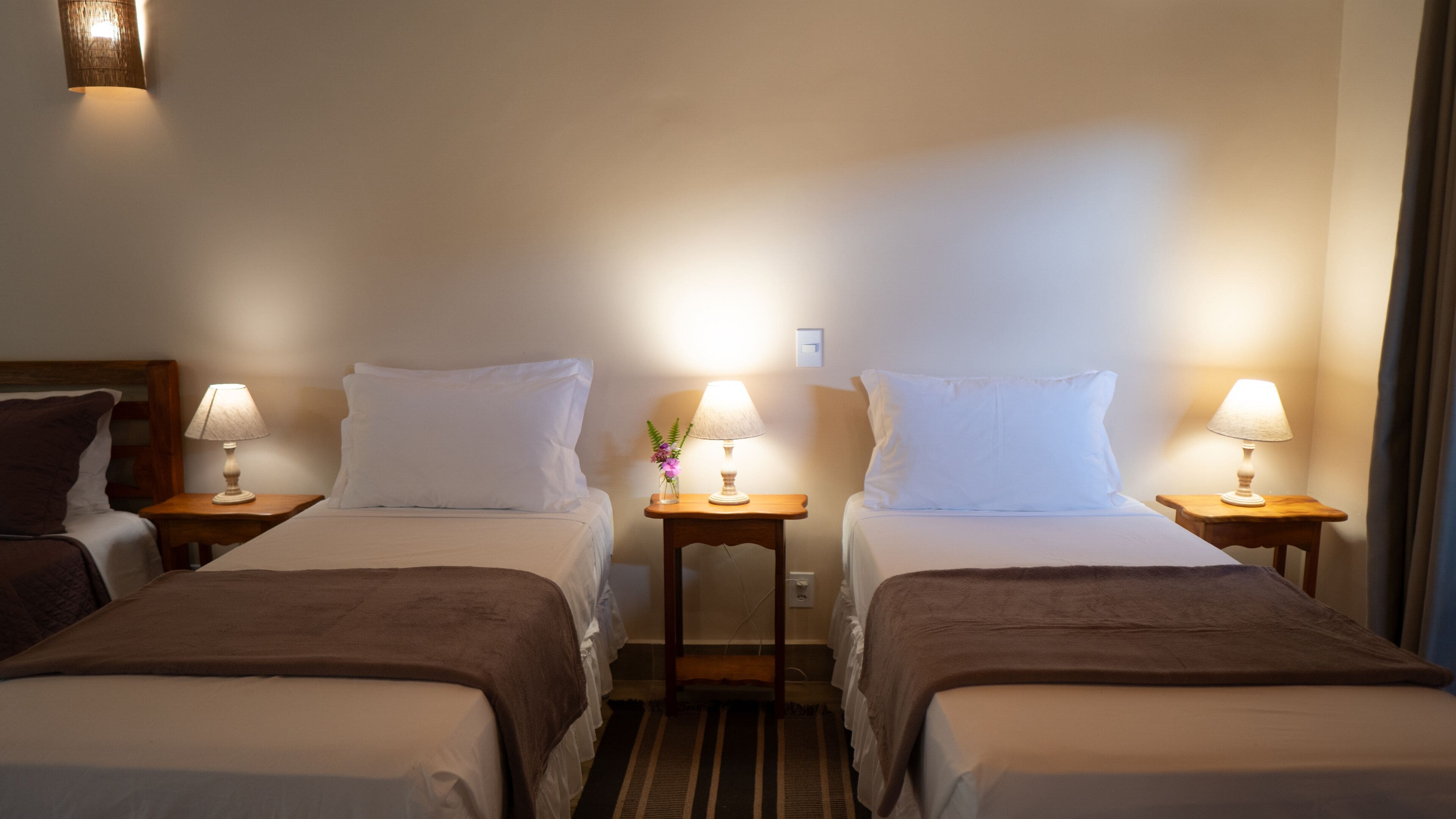 Camera d'albergo con due letti singoli, ognuno dotato di comodino e lampada. Un lato presenta una testiera in legno, l'altro una parete semplice.
