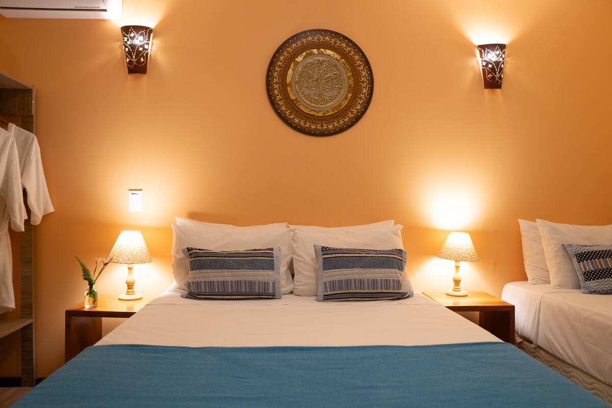 Quarto de hotel com cama, duas mesas de cabeceira, duas luminárias e um objeto decorativo na parede.