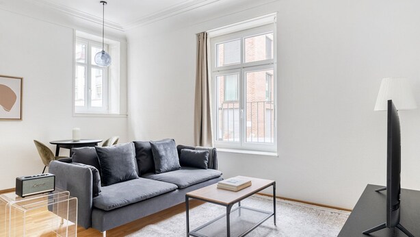 Ein modernes Wohnzimmer mit grauem Sofa, einem Couchtisch, einer TV-Konsole und einer Lampe. Zwei Fenster sorgen für natürliches Licht.