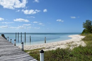 Un paseo marítimo de madera que conduce a una playa arenosa con agua azul cristalina y algunas nubes dispersas.