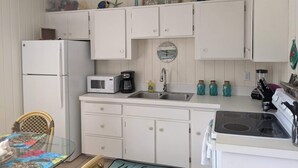 Une cuisine compacte avec des meubles blancs, un réfrigérateur, un four à micro-ondes, un évier et une cuisinière. On y trouve une chaise en osier et une table recouverte d’un joli tapis coloré.