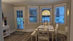 Un coin repas comprenant une table et des chaises, de grandes fenêtres équipées de stores, un luminaire suspendu et une petite table d’appoint agrémentée d’objets décoratifs.