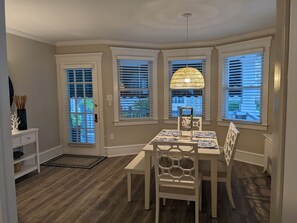 A dining area with a table and chairs, large windows with blinds, a hanging light fixture, and a small side table with decorative items.