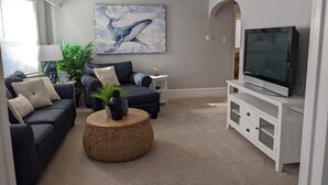 Una sala de estar con un sofá azul oscuro, una mesa de centro redonda, un televisor de pantalla plana sobre un mueble blanco y un ventilador de techo.
