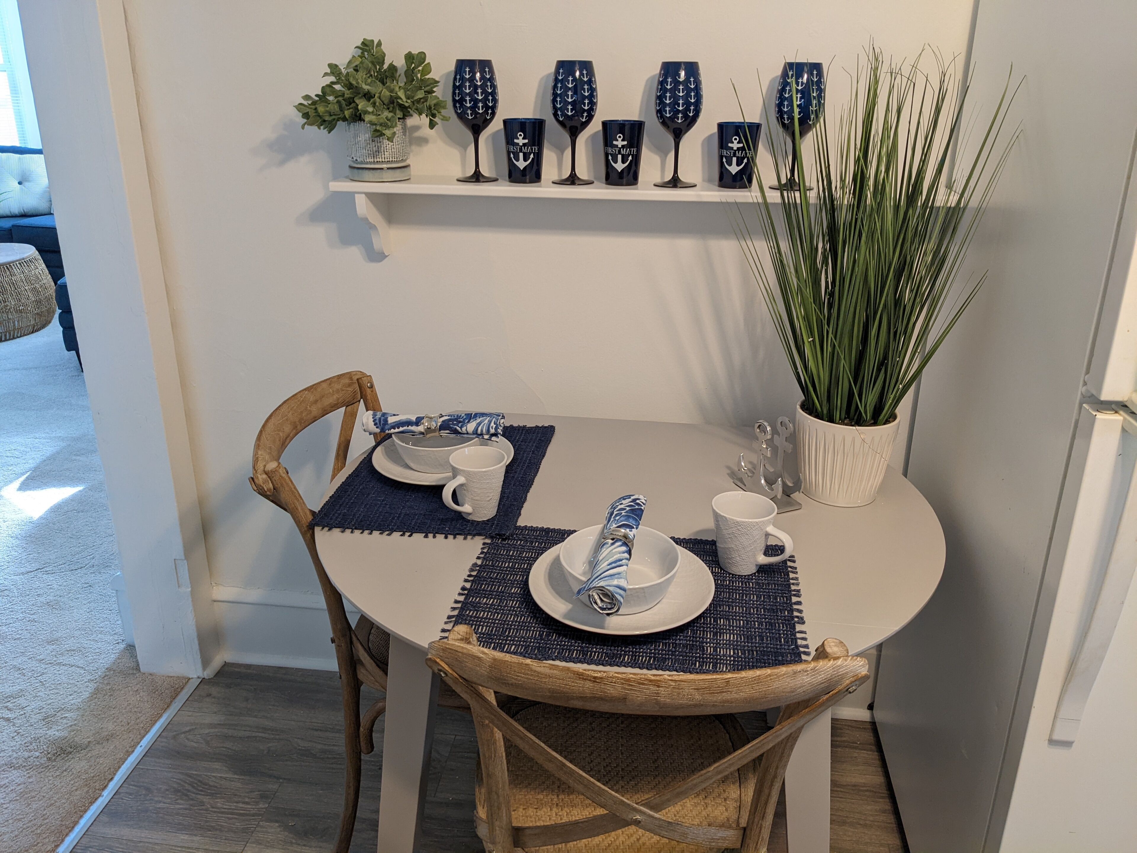 A dining table set with white plates, blue napkins, and a blue umbrella. There are six wine glasses with anchor designs on the wall above.