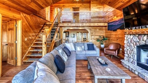 Un intérieur spacieux de chalet en rondins, avec un grand salon, une cheminée en pierre, un escalier en bois et un balcon.