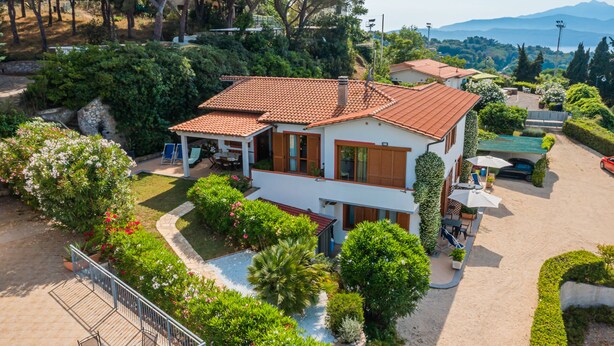 Une maison à deux étages, avec un toit recouvert de tuiles, entourée d’une végétation luxuriante et dotée d’une piscine.