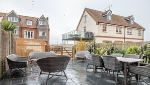 A patio area with wicker furniture and a stone floor, surrounded by a wooden fence and plants. - Blue Sky, Wells-next-the-Sea, Norfolk (Wells-next-the-Sea)