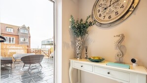 A room with a white console table, a wall clock, a vase with greenery, a decorative metal piece, and a bowl of fruit. - Blue Sky, Wells-next-the-Sea, Norfolk (Wells-next-the-Sea)